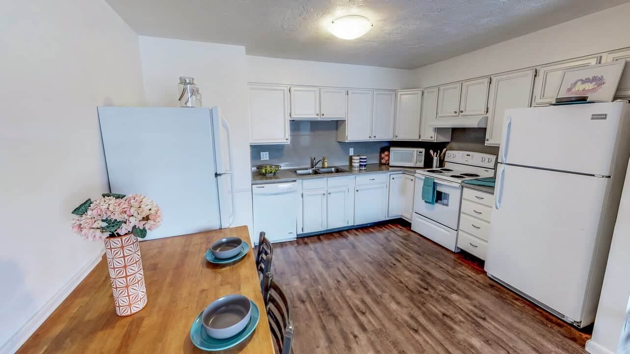 A bright kitchen at Somerset Apartments Rexburg features white cabinets, a white fridge, stove, and dishwasher. A wooden dining table with two place settings and pink flowers sits on a wood floor under overhead lighting.