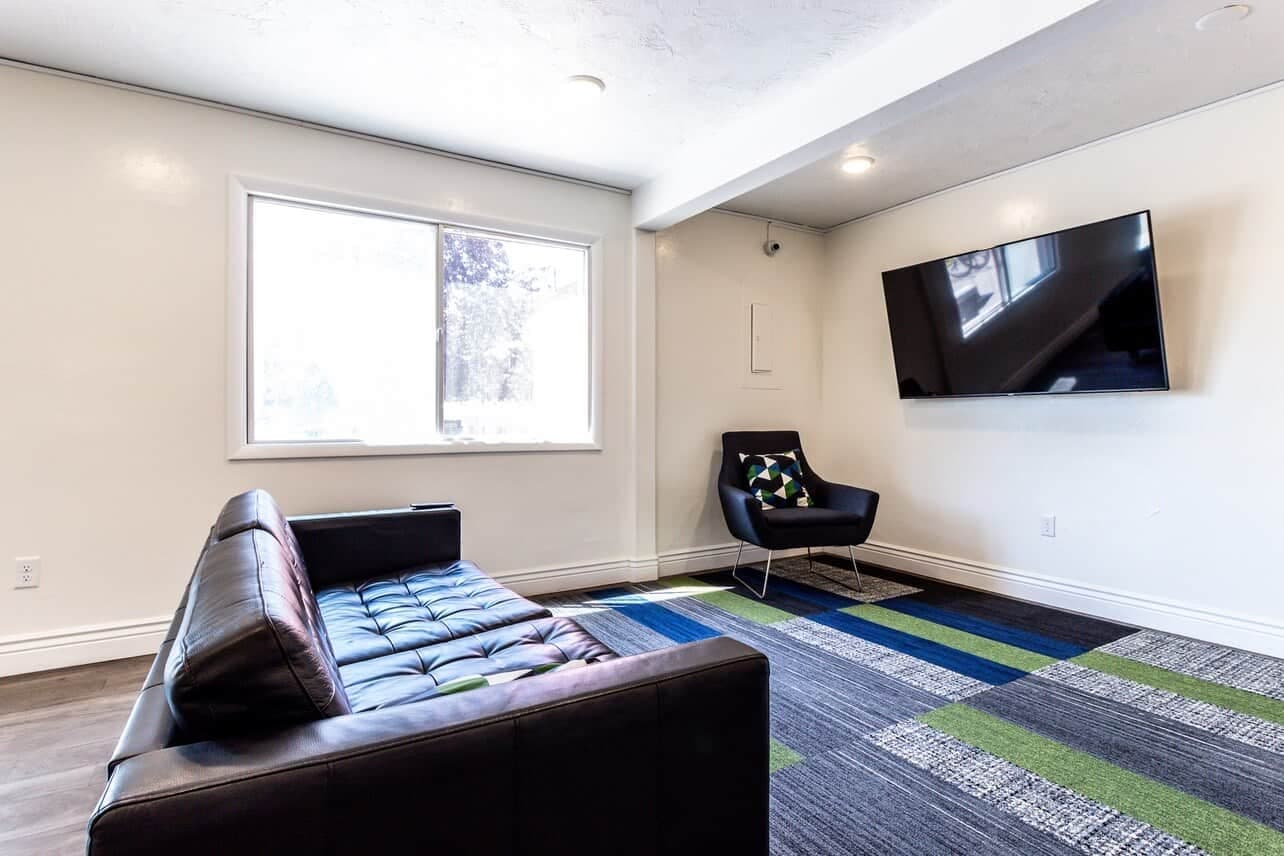 A modern living room at Somerset Apartments Rexburg features a black leather sofa, a black armchair with a patterned cushion, a wall-mounted TV, and natural light streaming in over a blue and green patterned rug.
