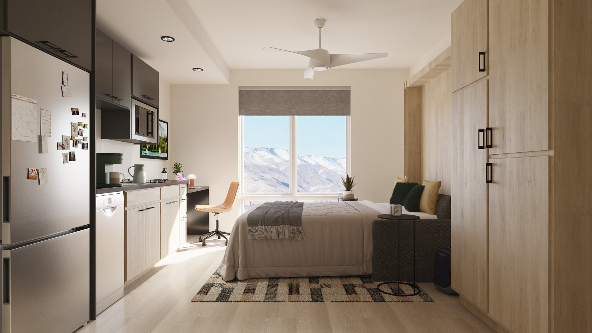 A modern studio apartment with a bed, desk, and chair by a window overlooking snowy mountains, a small kitchen area, light wood cabinets, a refrigerator, and a ceiling fan. The space is tidy and well-lit.