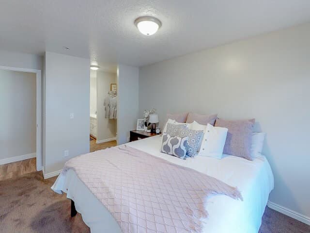 A neatly made bed with white and pink bedding sits in a cozy South Pointe Provo bedroom with beige walls and carpet. A nightstand with decor is beside the bed, and a doorway leads to a brightly lit closet area.