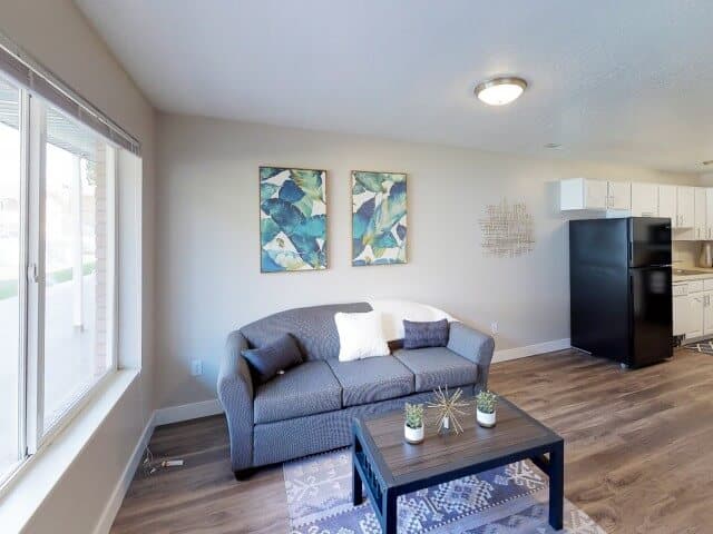 A cozy South Pointe Provo living room with a gray sofa, white and gray pillows, a coffee table with small plants, leaf-themed wall art, a large window, and part of a kitchen featuring a black refrigerator.