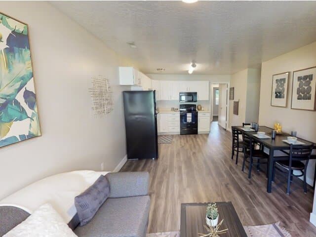 A modern open-concept living space at South Pointe Provo with wood flooring, a gray sofa, a small coffee table, a dining table for four, and a kitchen featuring white cabinets and black appliances. Abstract art decorates the walls.