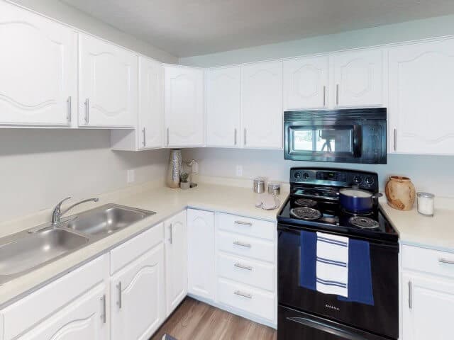 A bright South Pointe Provo kitchen with white cabinets, light countertops, a double sink, and a black stove with a microwave above. A pot sits on the stove, decor accents line the counters, and the floor is light wood.