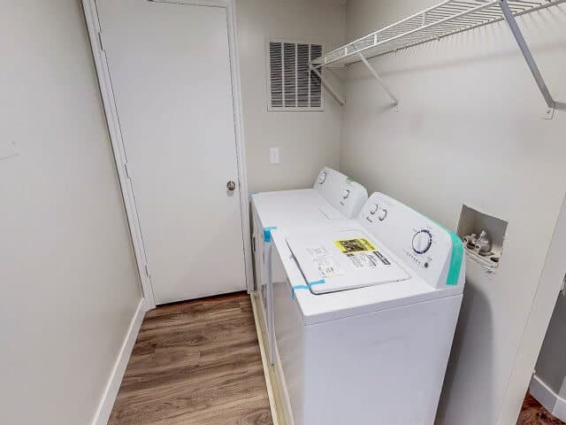 A laundry room at South Pointe Provo with a white washer and dryer side by side, a wire shelf above, closed door, vent, and exposed utility connections on the right wall. Light-colored walls and wood flooring complete the space.