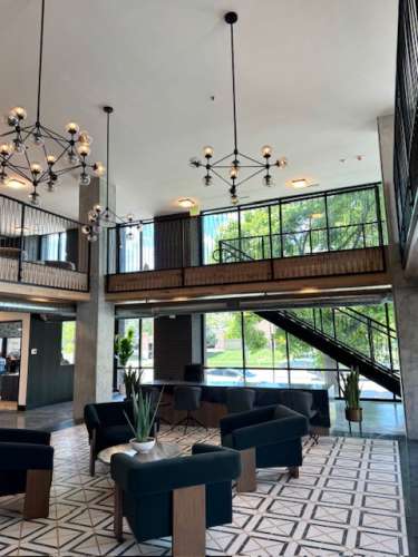 Modern hotel lobby at The Citizen Salt Lake City with geometric tile floor, navy blue chairs, potted plants, hanging chandeliers, and a staircase leading to an upper level with glass railings. Large windows reveal lush greenery outside.
