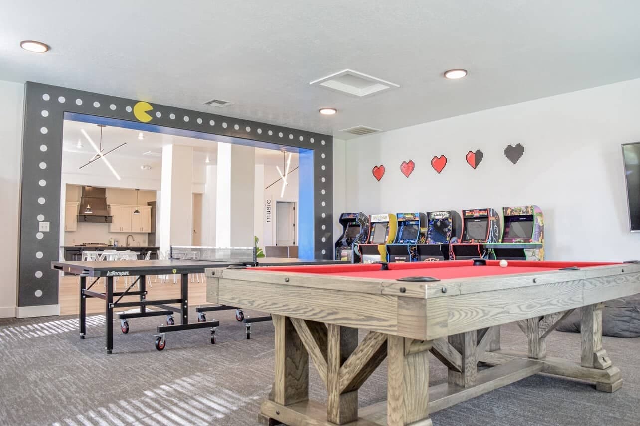 A game room at The Gates at Rexburg features a red pool table, a ping pong table, arcade machines, and Pac-Man themed decor on the wall. A stylish modern kitchen is visible in the background, making this Rexburg spot perfect for fun and relaxation.