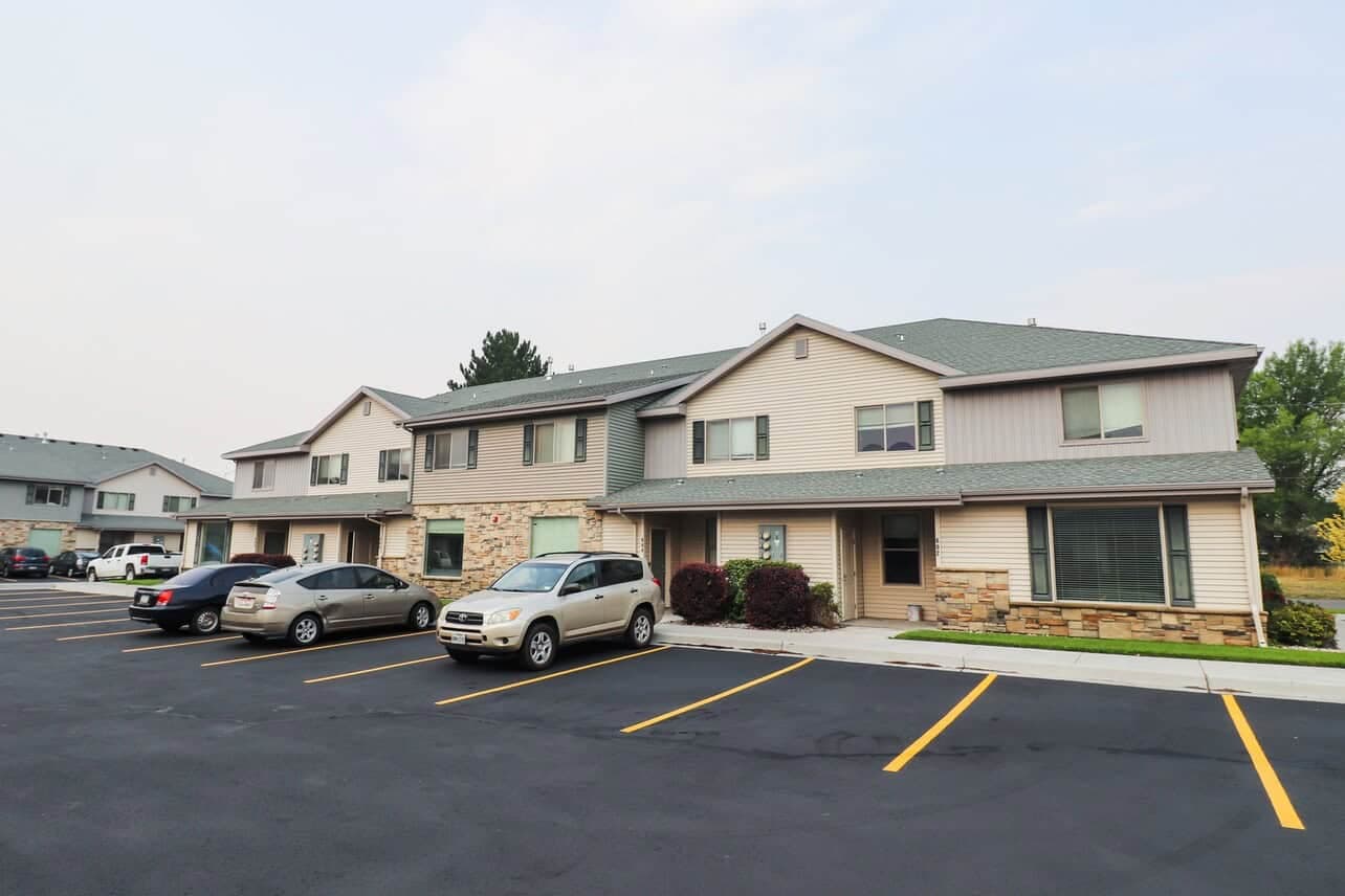 A row of modern two-story apartment buildings at The Gates at Rexburg with beige siding and stone accents, parked cars in a freshly-paved lot with yellow lines, and some greenery in the background under a light sky in Rexburg.