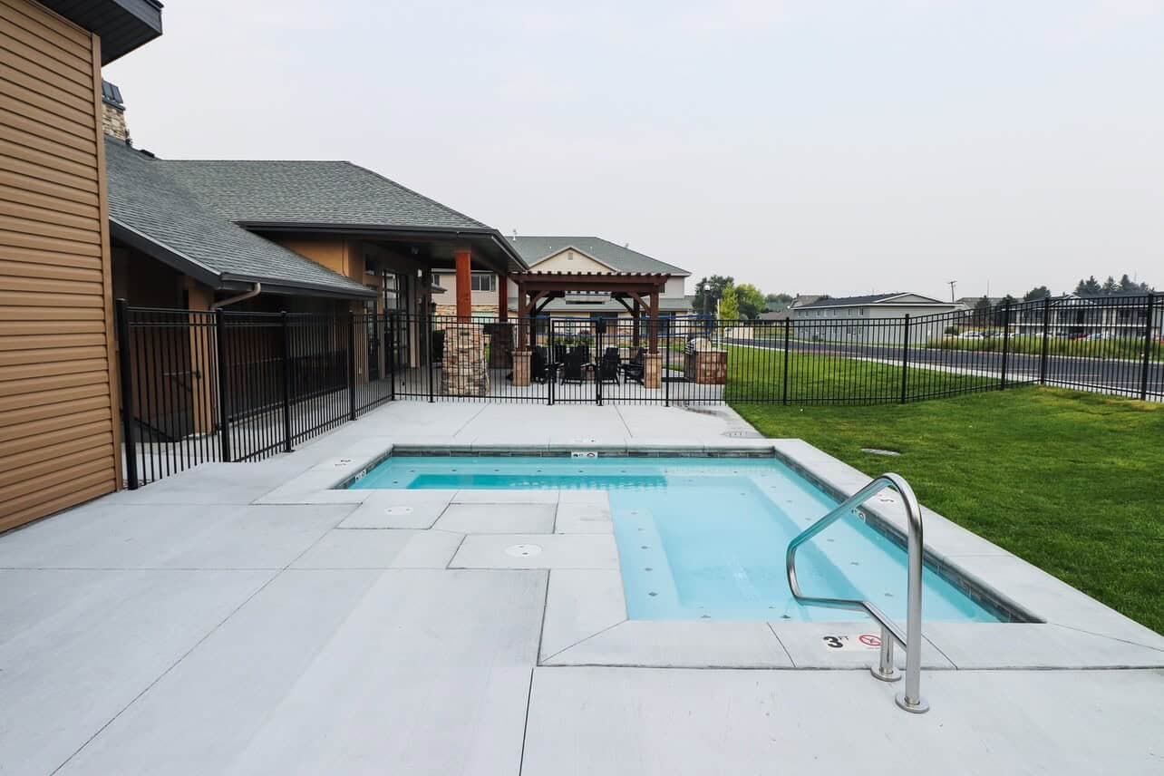 Outdoor view of a small rectangular pool and hot tub at The Gates at Rexburg, surrounded by a concrete deck, safety fence, and green grass, with a building and covered seating area in the background.