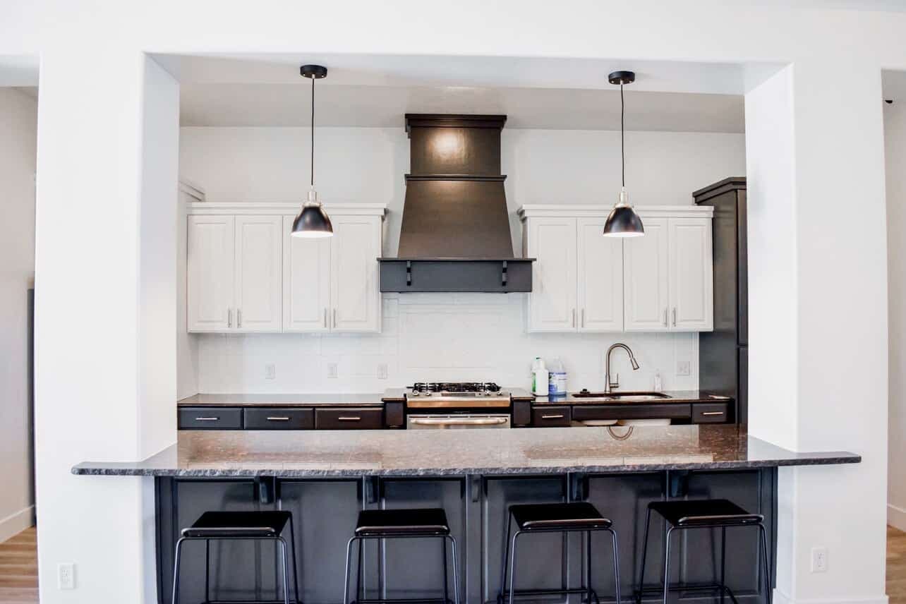 Modern kitchen at The Gates at Rexburg with white upper cabinets, dark lower cabinets, a black range hood, stainless steel appliances, a large island with a dark countertop, and four black bar stools under pendant lights.