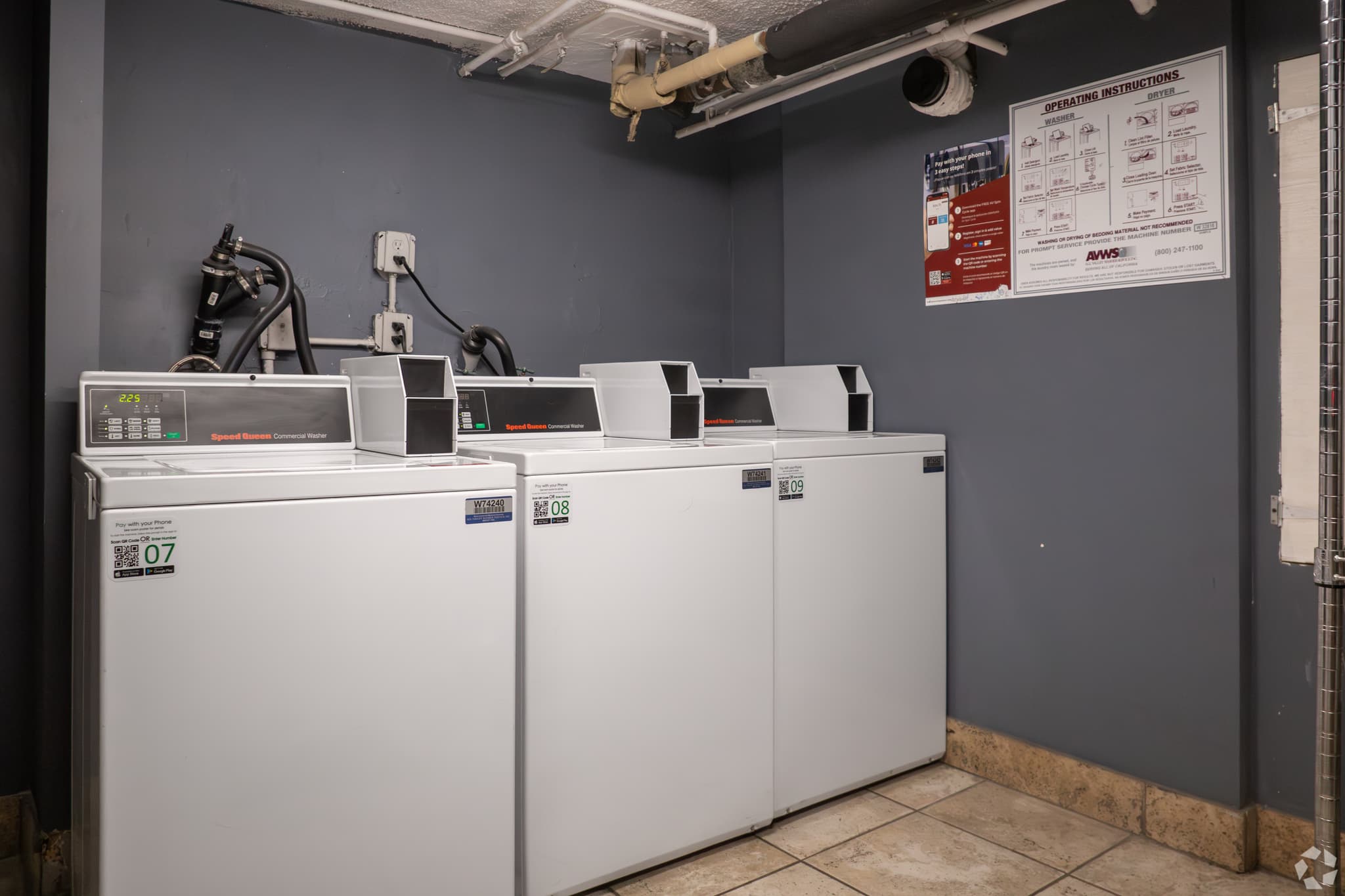 A small laundry room at Hive Ellendale features three white washing machines lined up against a gray wall. Instructions are posted above the machines, and utility pipes run near the ceiling.