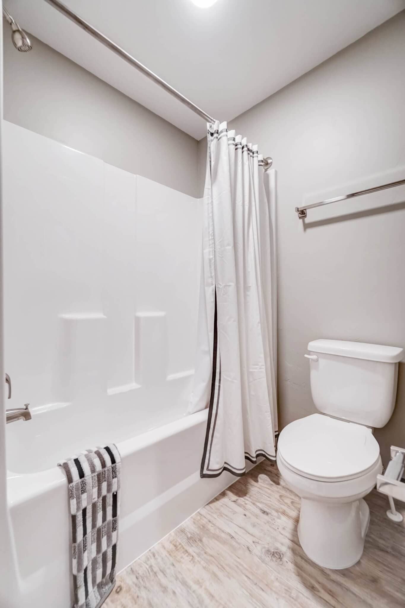 A clean, modern bathroom at The Landing Rexburg features a white bathtub and shower with a white curtain, a striped towel on the tub, a toilet, towel rack, light wood-style flooring, and light beige walls.