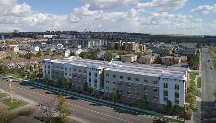 The Landing Rexburg is a wide, modern three-story apartment building situated along a main road in a suburban neighborhood, surrounded by rows of houses, trees, and open sky.