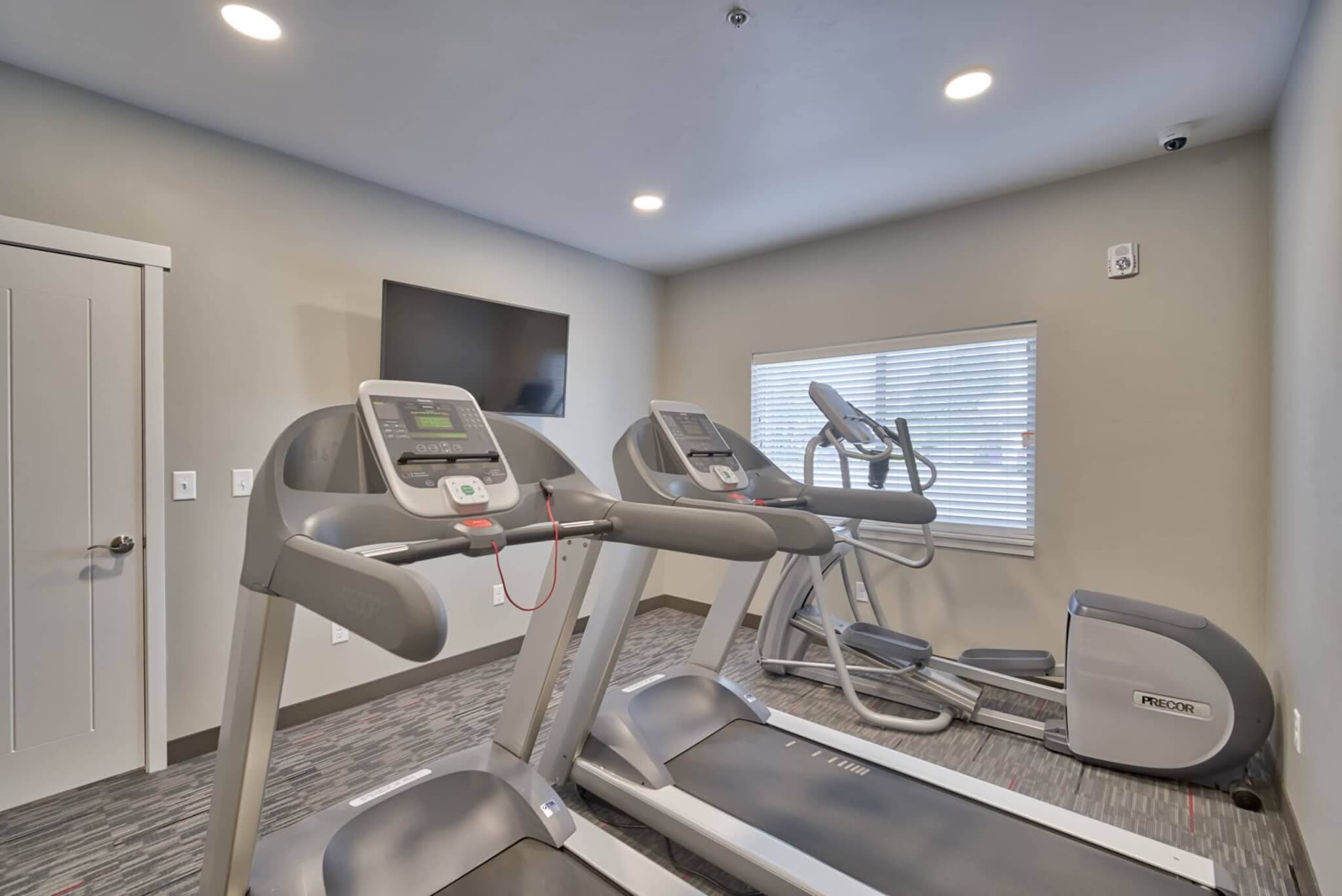 A small gym room at The Landing Rexburg features two treadmills, an elliptical machine, a stationary bike, a wall-mounted TV, and a window with blinds. The space is finished with gray carpet and light-colored walls.