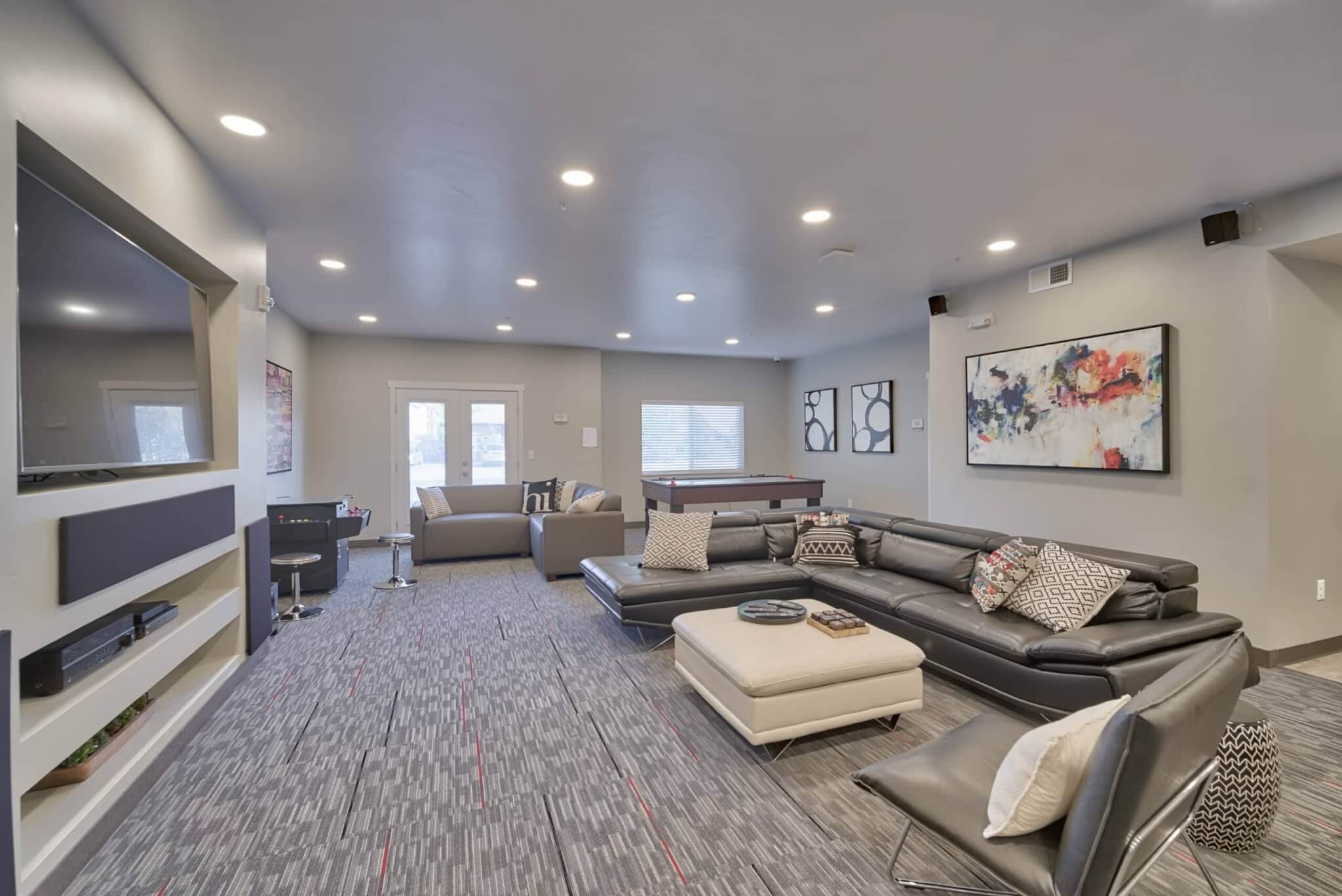 Modern living room at The Landing Rexburg with gray walls, recessed lighting, a large black sectional sofa, ottoman, accent chair, wall art, TV, and a pool table. Windows let in natural light for an open and inviting space.