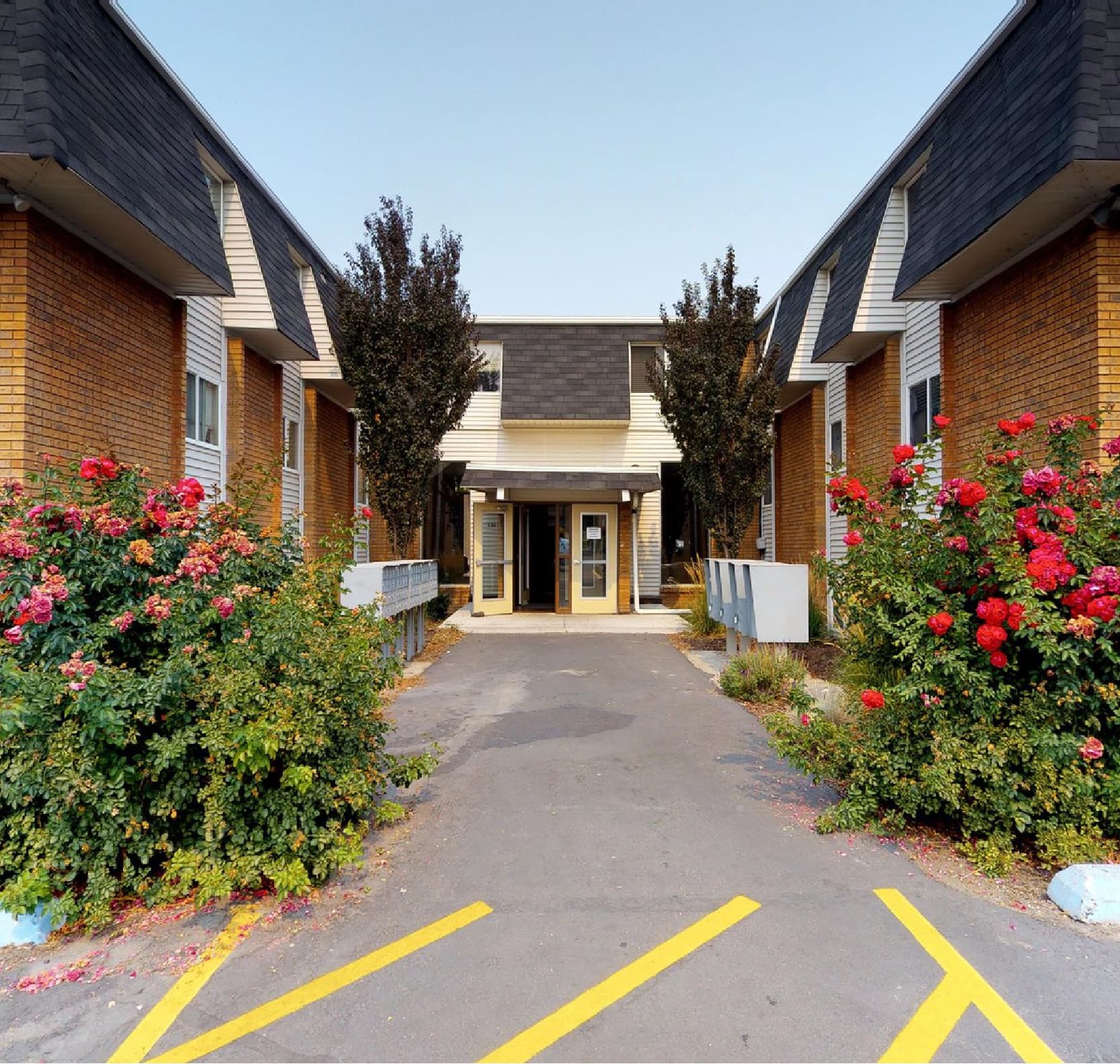 The Pines Rexburg is a two-story brick building with a central entrance, flanked by bushes with red flowers and yellow painted lines on the asphalt in the foreground.