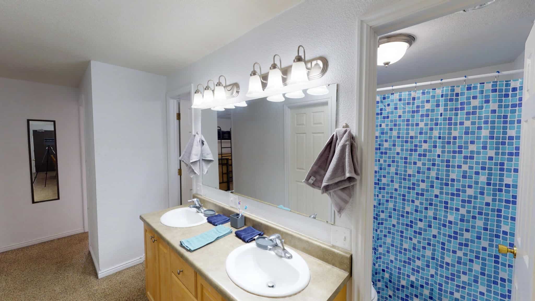 Modern bathroom at The Pines Rexburg with double sinks, a large mirror, towels, and blue mosaic shower curtain. The countertop holds towels and soap dispensers; bright lights above the mirror illuminate the space.