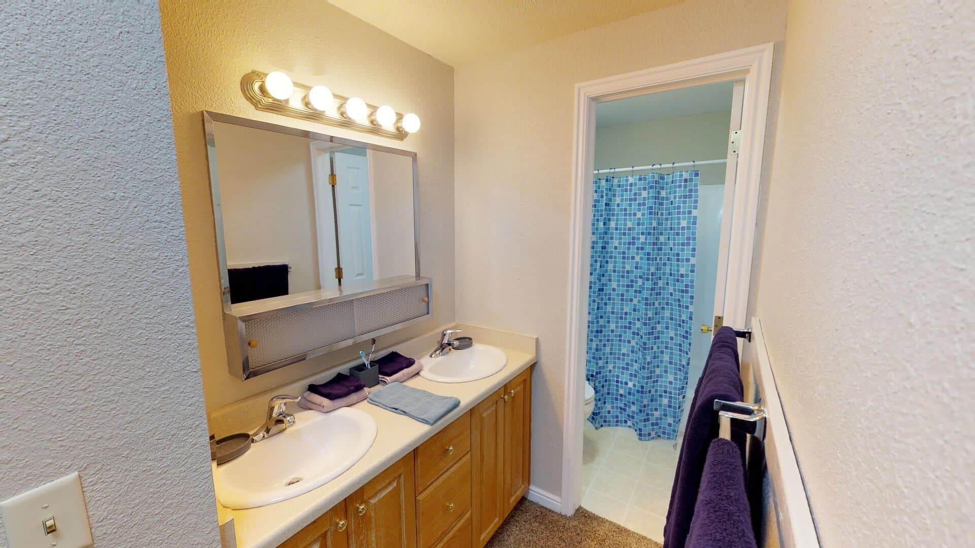 A bathroom at The Pines Rexburg features a double sink vanity, large mirror, wall lights, and purple towels. In the background, a shower with a blue mosaic-patterned curtain stands over a pristine white tile floor.
