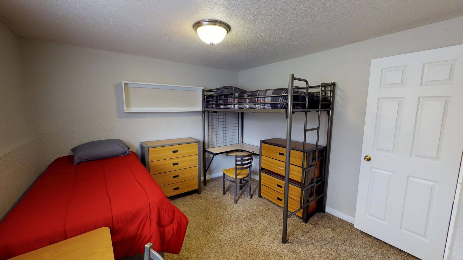 A cozy small bedroom at The Pines Rexburg features a single bed with a red blanket, a lofted bunk bed with a desk and chair underneath, two wooden dressers, beige carpet, and a white door.