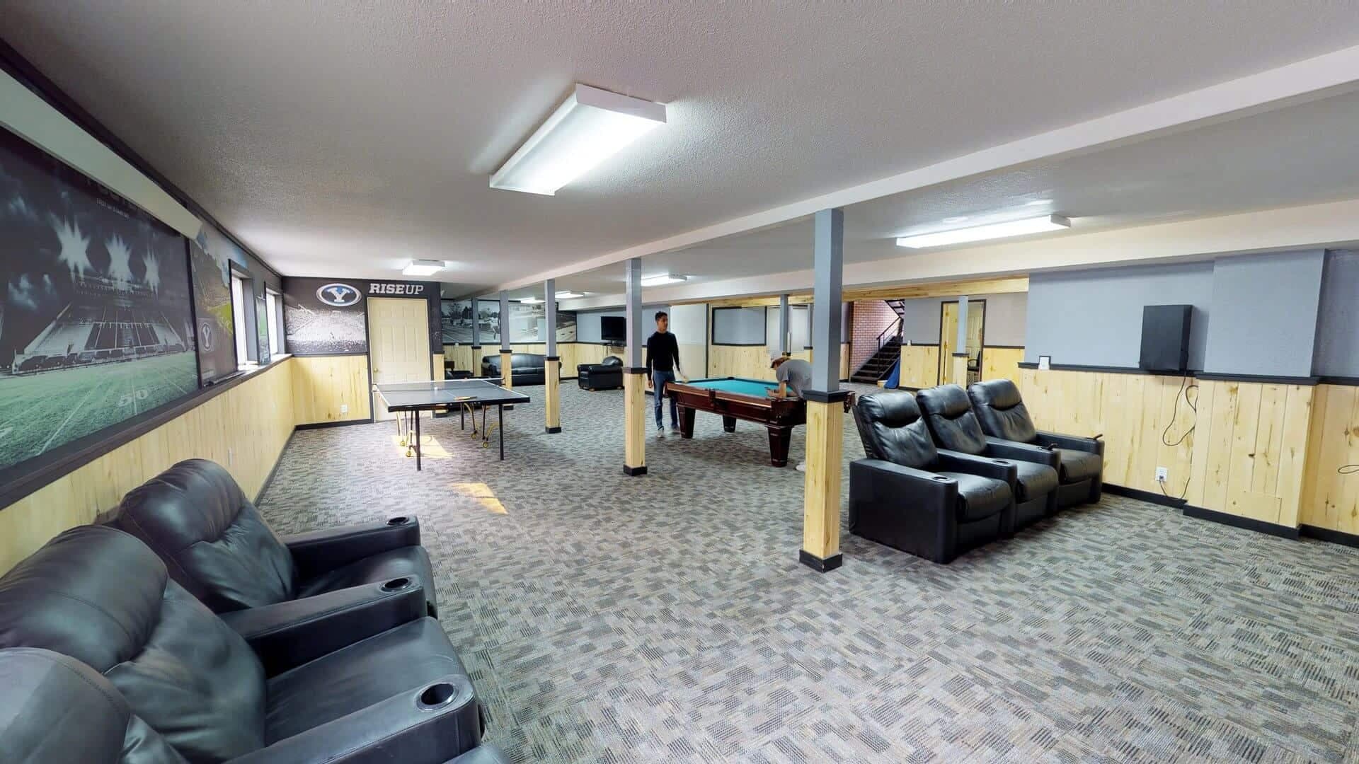Spacious game room at The Pines Rexburg with recliner seats, pool table, ping pong table, wall art, and two people playing pool. The room features carpeted floors, wood paneling, and bright overhead lighting.