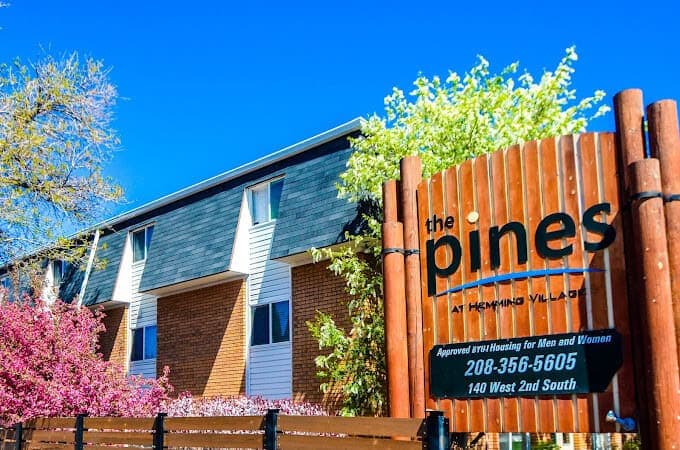 A brick apartment building with white trim sits behind a wooden fence at The Pines Rexburg. A sign reads The Pines at Hemming Village, Approved BYU-I Housing for Men and Women, with a phone number and address. Trees bloom nearby.