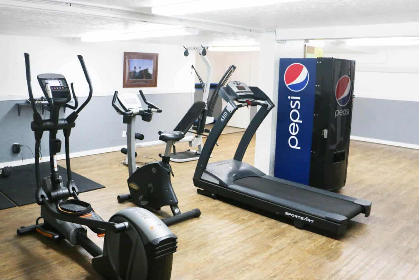 A small gym room at The Pines Rexburg features exercise equipment including a treadmill, stationary bikes, an elliptical machine, and a large Pepsi vending machine, all set on a wood floor with white walls.