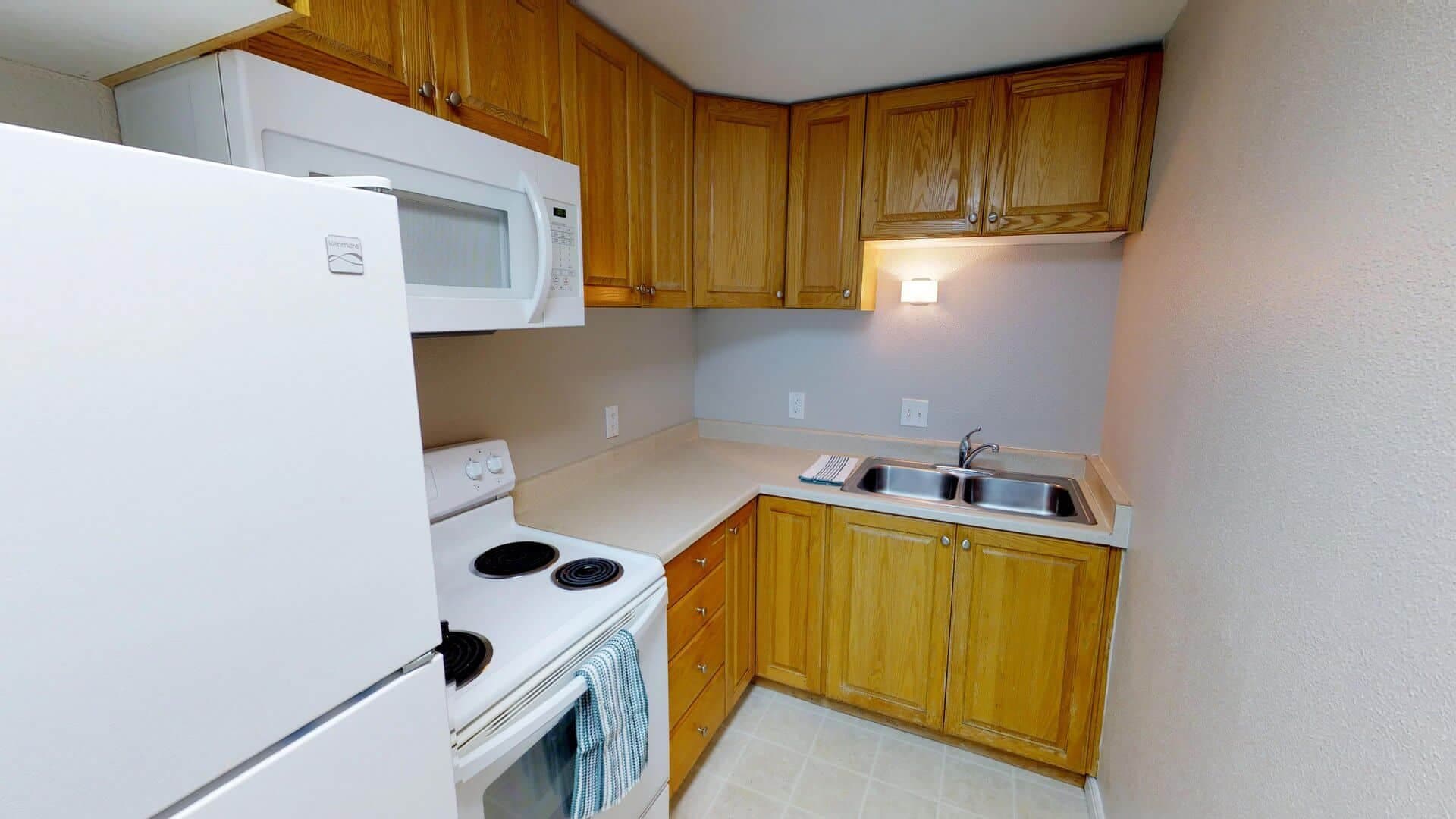 A small kitchen at The Pines Rexburg features wooden cabinets, white countertops, a white refrigerator, a stove with a towel, a microwave, a stainless steel sink, and beige walls. The space is clean and well-lit.