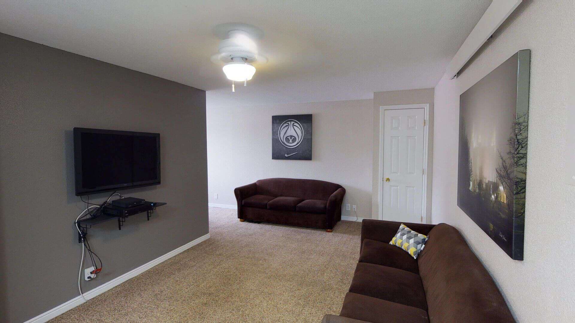 A cozy living room at The Pines Rexburg features brown sofas, a wall-mounted TV, a ceiling fan, beige carpet, and framed art on light-colored walls. A door sits in the background beside a small shelf under the TV.