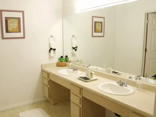A clean bathroom at The Ridge Rexburg features a long countertop with two sinks, a large mirror, and light wood cabinets. Framed art pieces decorate the wall, along with soap dispensers, green bottles, and a white rug on the floor.