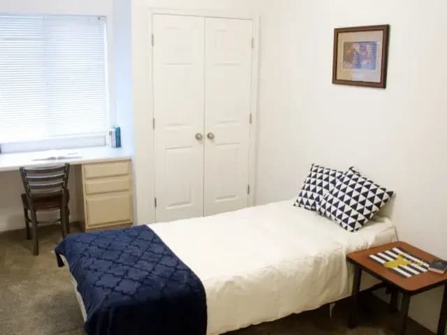 A simple bedroom at The Ridge Rexburg features a single bed with a blue blanket, geometric pillows, a small table with a few items, a desk and chair by the window, a double-door closet, and a framed picture on the white wall.
