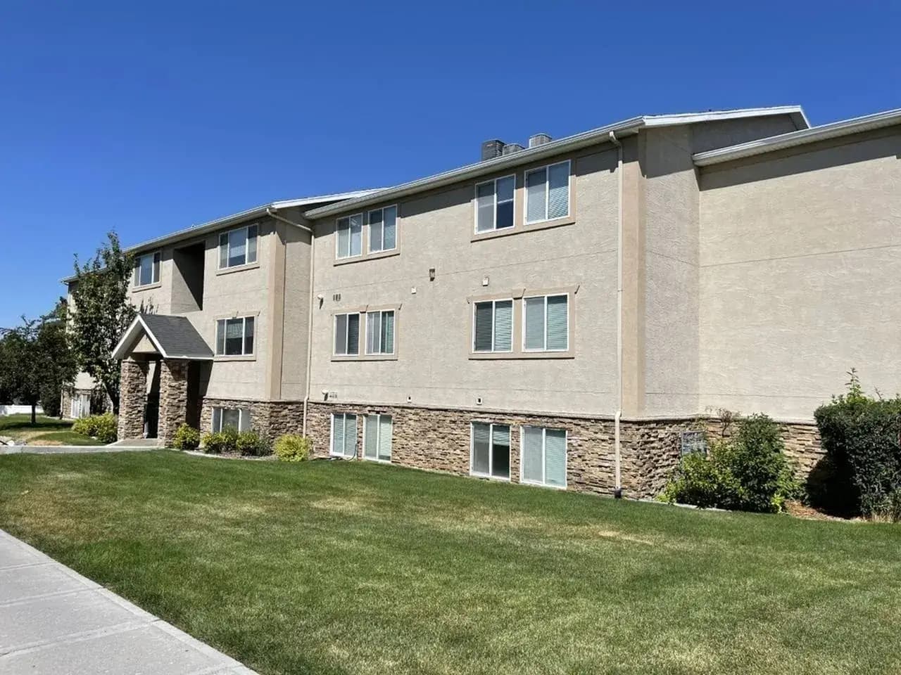The Ridge Rexburg: A beige, two-story apartment building with stone accents, multiple windows, and a green lawn in front under a clear blue sky. Shrubs line The Ridge Rexburg and a sidewalk runs along the left side.
