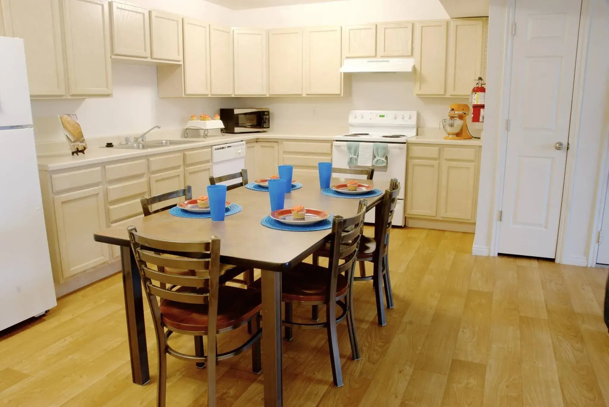 A kitchen at The Ridge Rexburg features light wood cabinets, white appliances, and a wooden dining table set for four with blue plates and cups. The room has a light wood floor and a visible fire extinguisher on the wall.