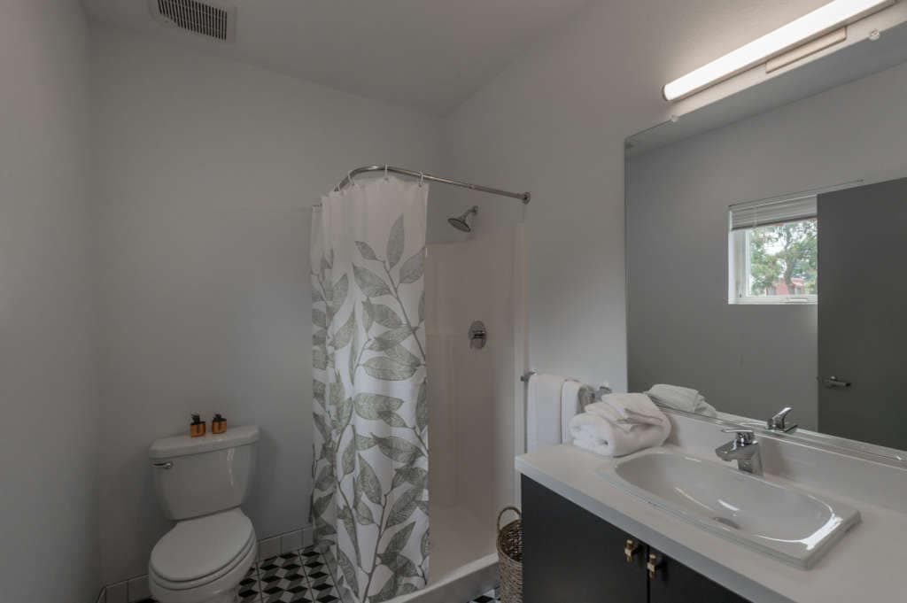 A bathroom at The Urban Chico, featuring a toilet, a shower with a leaf-patterned curtain, a sink with mirror above, toiletries, towels, and a small window letting in natural light.