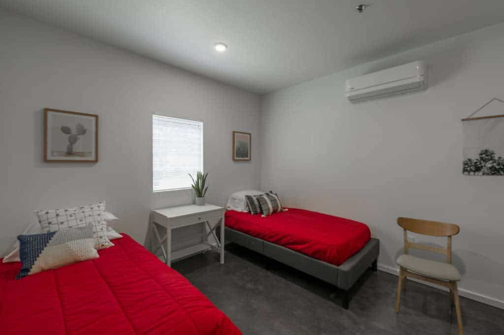 A clean, modern bedroom at The Urban Chico features two single beds with red bedding, a small white desk with a plant, a wooden chair, wall art, and an air conditioning unit above. Light enters through a window between the beds.