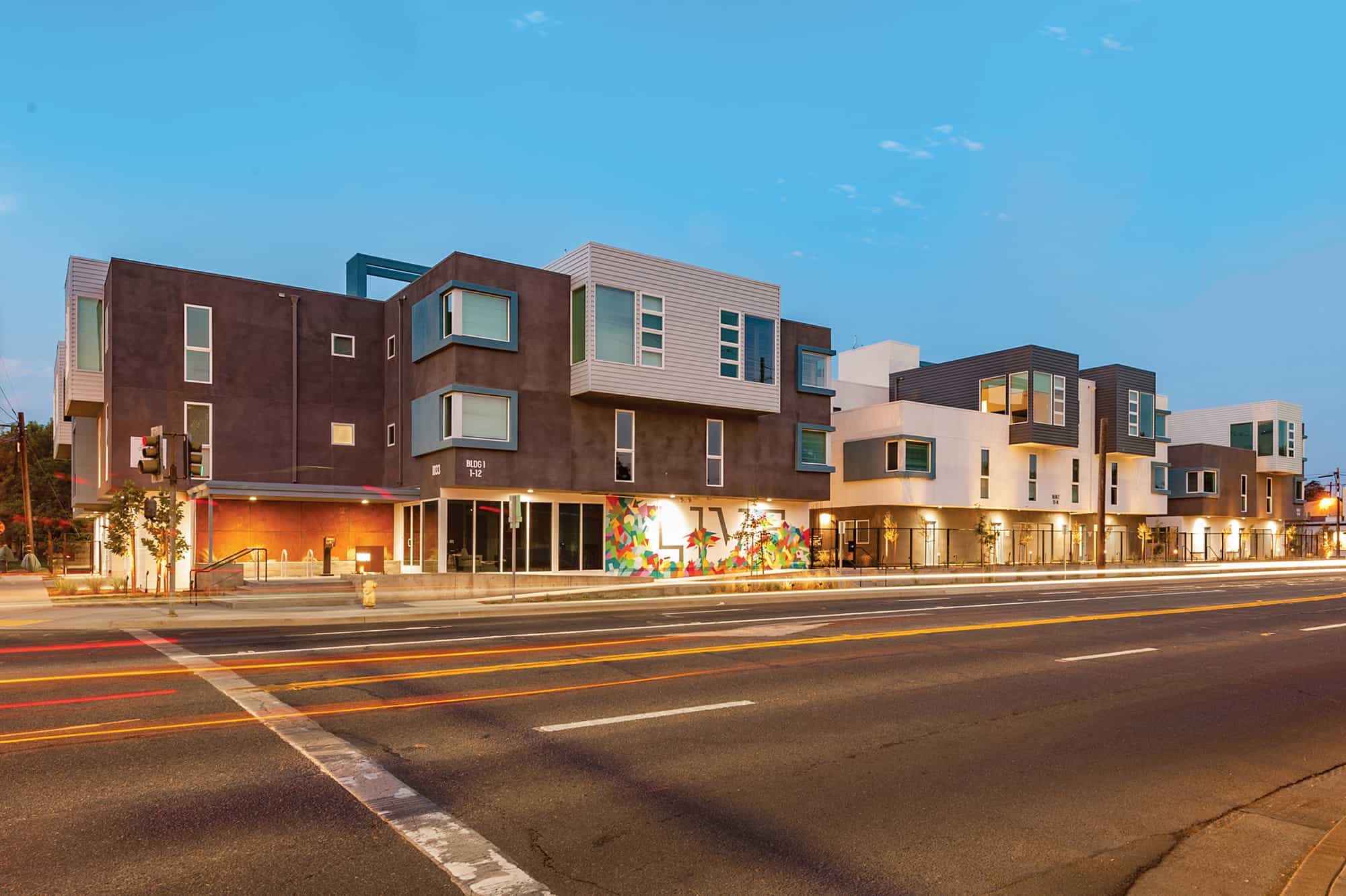 Modern apartment complex, The Urban Chico, features boxy architecture, large windows, and a colorful mural on the lower level, illuminated at dusk with blurred car lights streaming by in front.