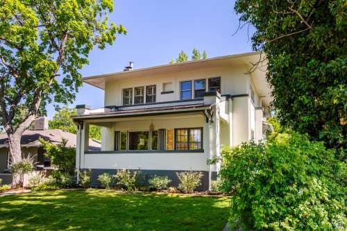 The Utah Prairie House Salt Lake City is a two-story white home with a covered front porch, surrounded by green trees and bushes beneath a clear blue sky. A well-maintained lawn stretches across the front yard.