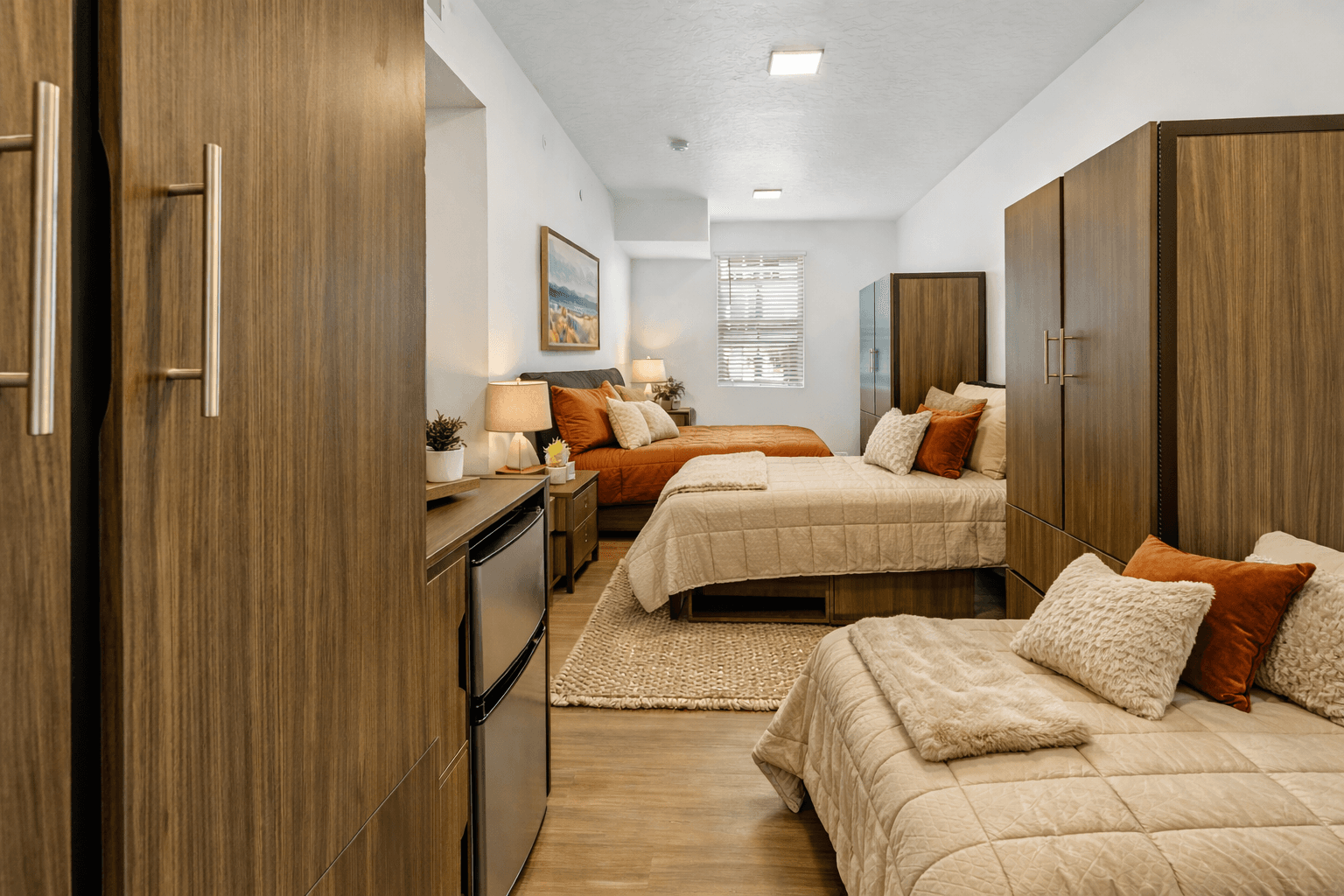 A modern dorm room at Slopeside Village with three beds, neutral bedding, wood furniture, a mini fridge, window blinds, wall art, bedside lamps, and potted plants—creating a cozy shared living space near slopeside lodging in Park City.