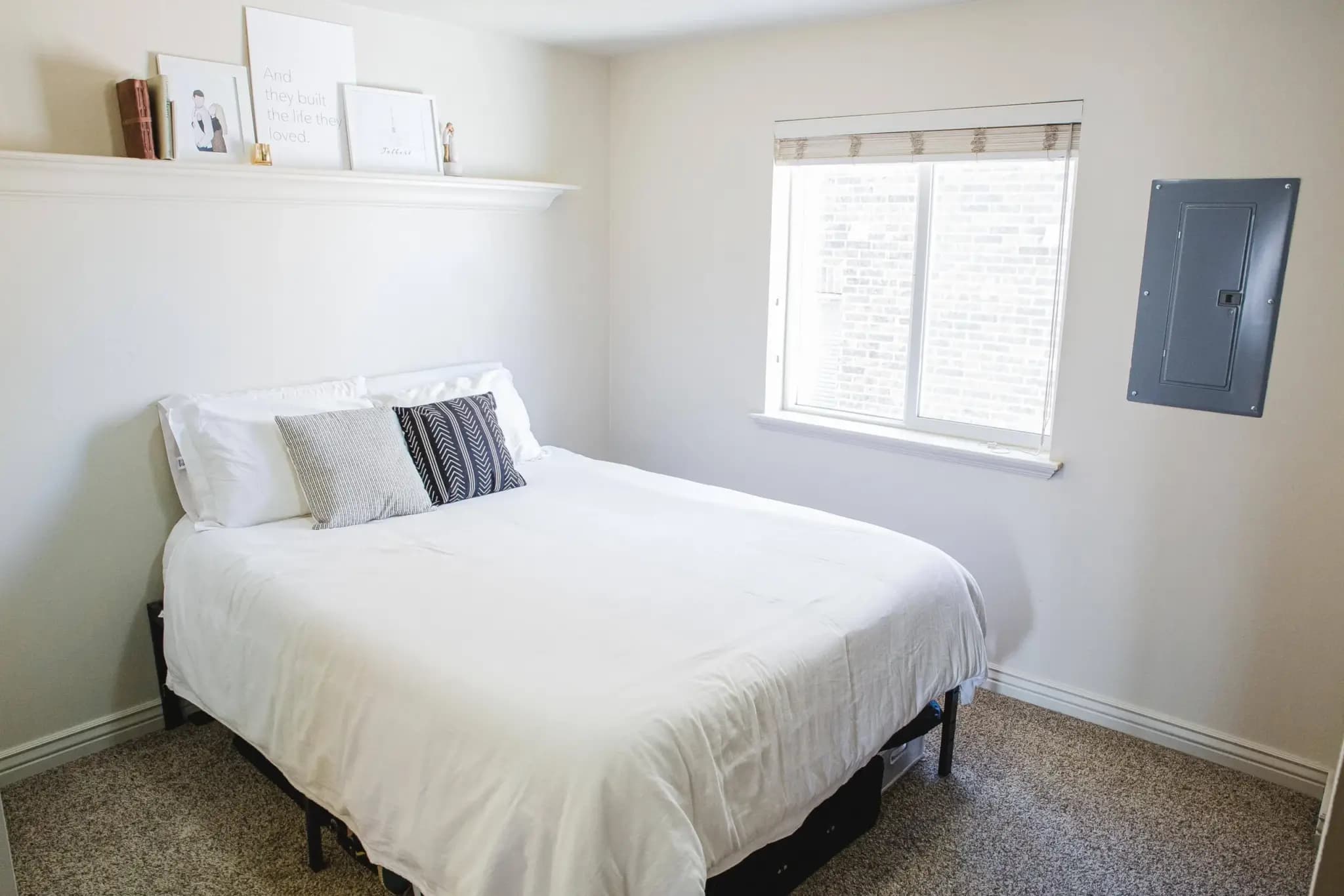 A small, bright University Courtyard Rexburg bedroom with a neatly made bed covered in white bedding and accent pillows, a shelf with decor above the bed, a window with blinds, and a gray electrical panel on the wall.