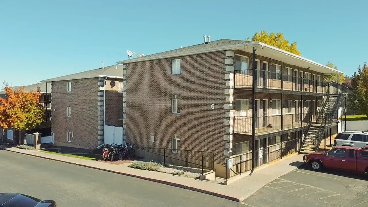A three-story brick apartment building at University Courtyard Rexburg features exterior staircases and balconies on a sunny day. Several parked cars, bicycles near the entrance, and autumn-colored trees complete the scene.