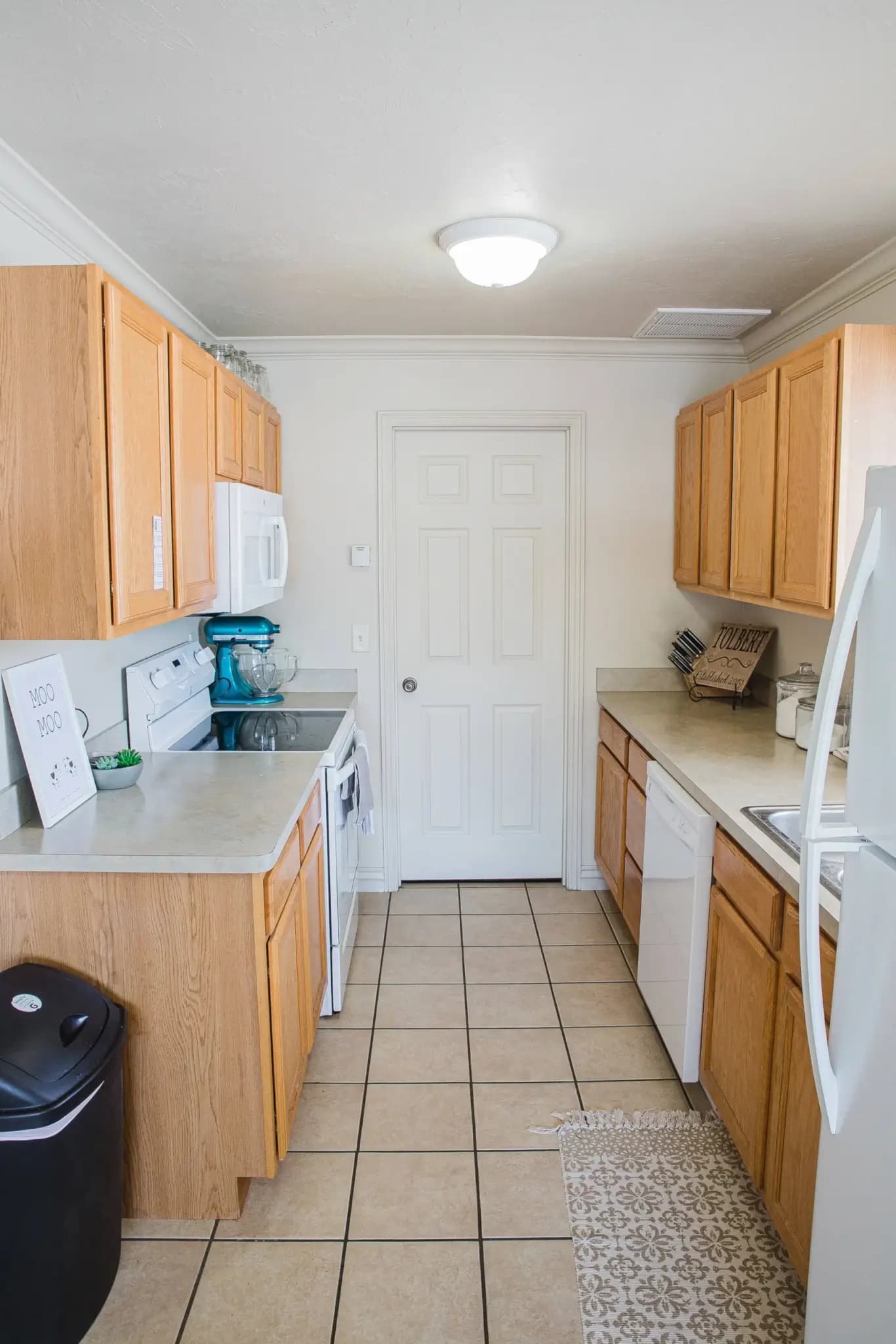 A bright, narrow kitchen at University Courtyard Rexburg features light wood cabinets, white appliances, beige countertops, and a tiled floor. A door is at the far end, with a patterned rug and small dish rack by the sink.