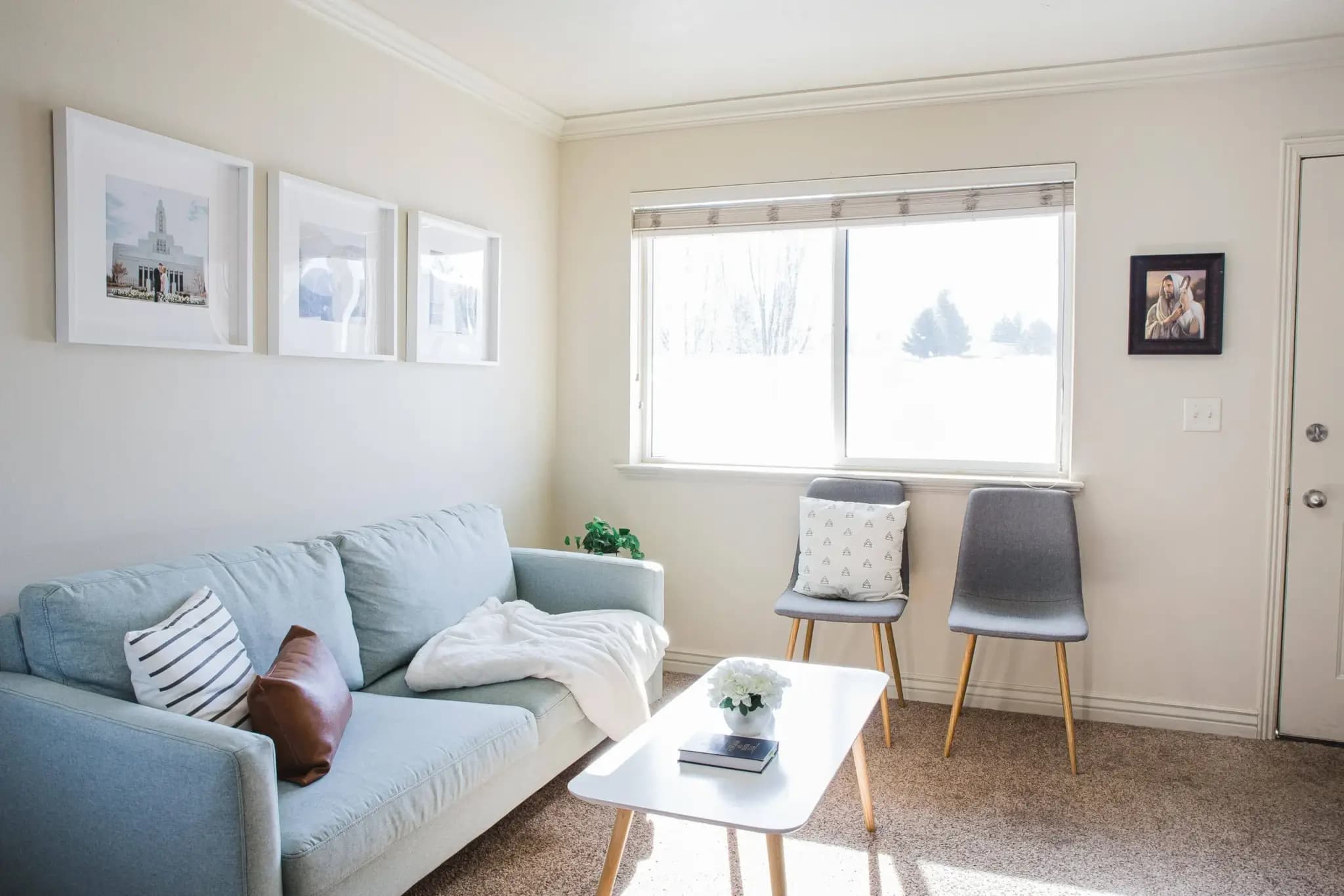 Bright living room at University Courtyard Rexburg with a light blue sofa, two gray chairs, a white coffee table, framed wall art, sunlight from the window, and neutral-colored decor for a cozy, minimalist feel.
