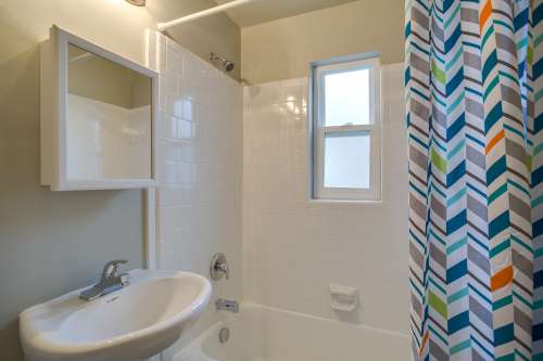 Small bathroom at University Gardens Apartments Salt Lake City with white tile walls, a white sink, bathtub, wall-mounted mirror cabinet, window, and a shower curtain featuring a colorful chevron pattern.