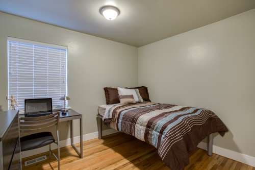 A small, tidy bedroom at University Gardens Apartments in Salt Lake City features a single bed with brown, blue, and white striped bedding. A simple desk and chair sit by the window with closed blinds, illuminated by a ceiling light.