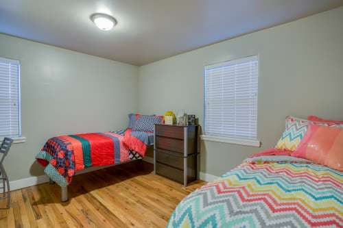 Bedroom with two single beds, colorful patterned bedding, and a dark wood dresser, in the inviting atmosphere of University Gardens Apartments Salt Lake City. Two windows with white blinds and wooden floors add to the bright, modern feel.