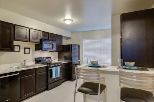 Modern kitchen at University Gardens Apartments Salt Lake City features dark wood cabinets, black appliances, light countertops, and a breakfast bar set for two. A window with blinds fills the space with natural light.