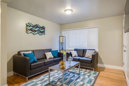 Modern living room at University Gardens Apartments Salt Lake City with two dark sofas, turquoise and white cushions, geometric rug, coffee table, floor lamp, wall art, and a window with blinds letting in natural light.