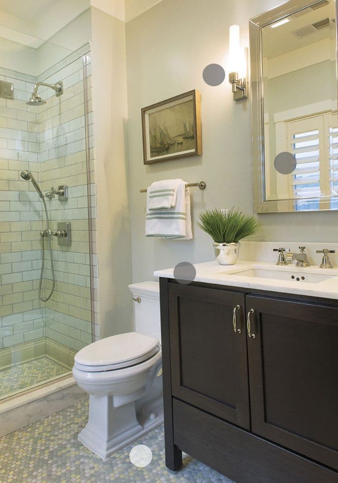 Modern bathroom with glass shower, white toilet, dark wood vanity, white sink, wall-mounted towel bar with towels, framed artwork, potted plant, and light fixture above a mirror on a light-colored wall.