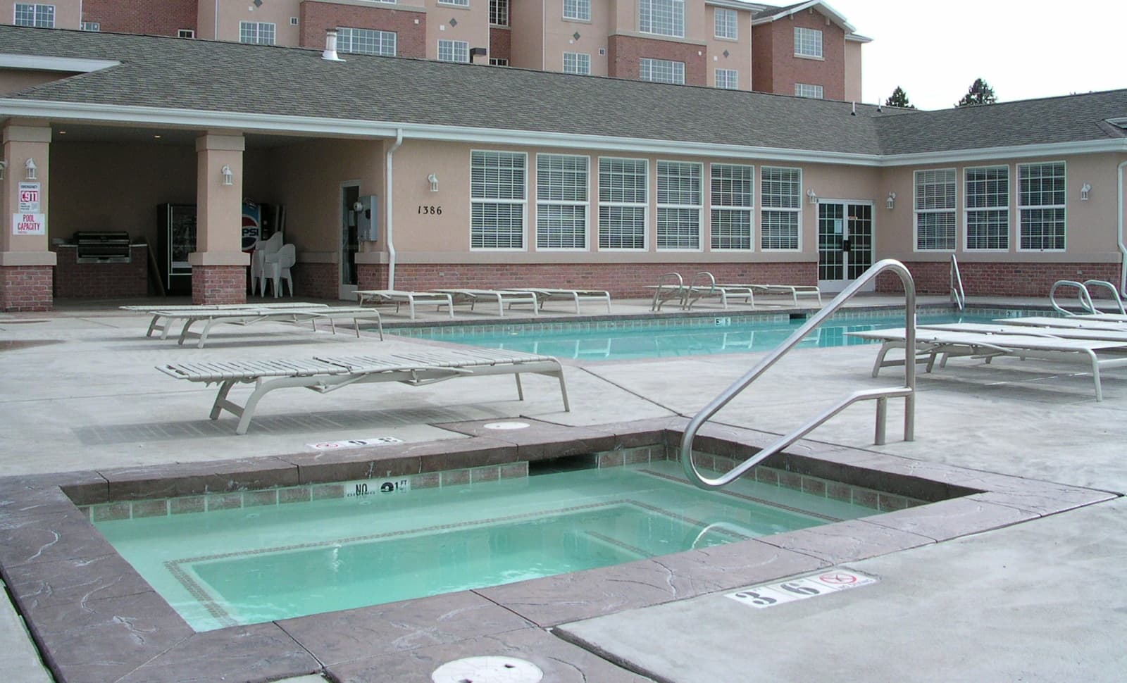 Outdoor pool area with a hot tub in the foreground, several lounge chairs, and a larger swimming pool behind it. The building in the background has many windows and a covered patio with seating.