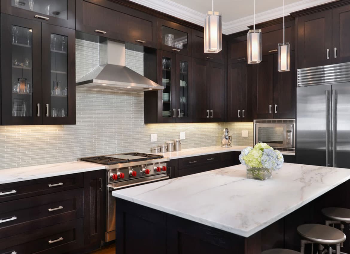 Modern kitchen with dark wood cabinets, stainless steel appliances, white marble countertops, a central island with a vase of flowers, glass tile backsplash, and pendant lights hanging from the ceiling.