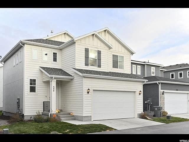 Two-story modern suburban house with light-colored siding, a double garage, and a small front yard with minimal landscaping. The house is situated between similar neighboring homes under a partly cloudy sky.