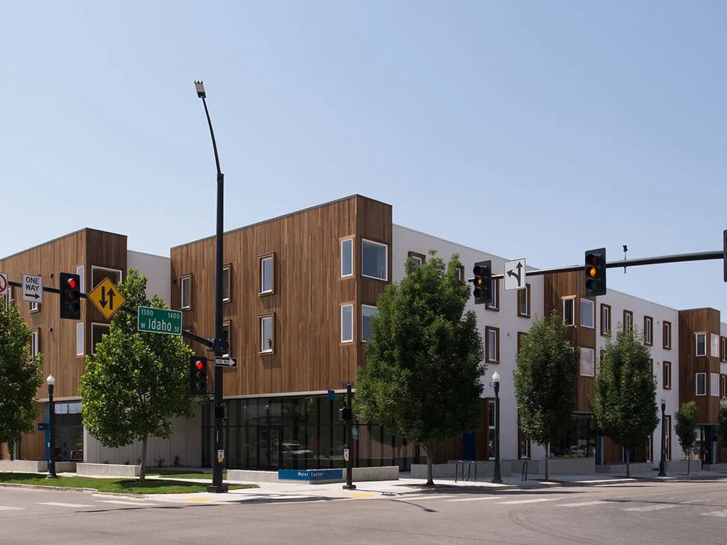 A modern, three-story building named Watercooler Apartments with a wood and white exterior sits at a street corner with traffic lights and green trees. The street signs read Idaho and 9th St. The sky is clear and blue.